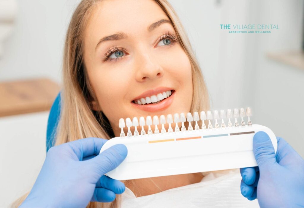 Woman in dental chair smiling while comparing tooth shade guide for porcelain veneers at The Village Dental in Pinecrest, FL, highlighting cosmetic dentistry services for smile makeovers.