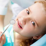 Smiling young girl at The Village Dental in Pinecrest, FL, holding a dental mirror, showcasing a positive and comfortable dental visit experience.