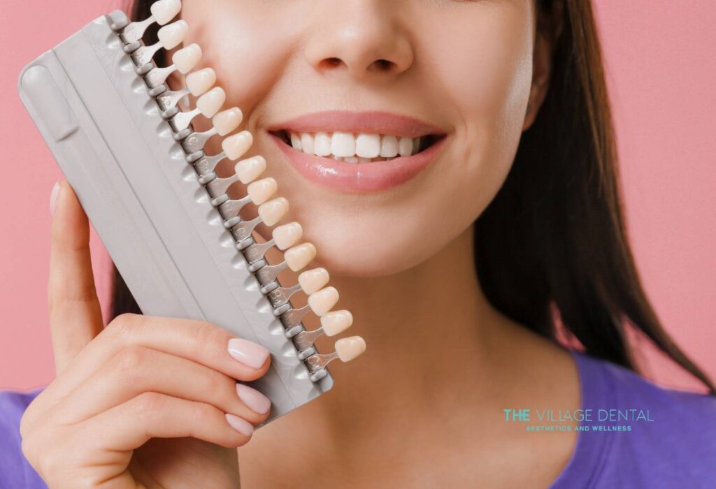Woman smiling while holding a dental shade guide next to her teeth, showcasing options for porcelain veneers at The Village Dental in Pinecrest, FL, emphasizing cosmetic dentistry and smile enhancement.