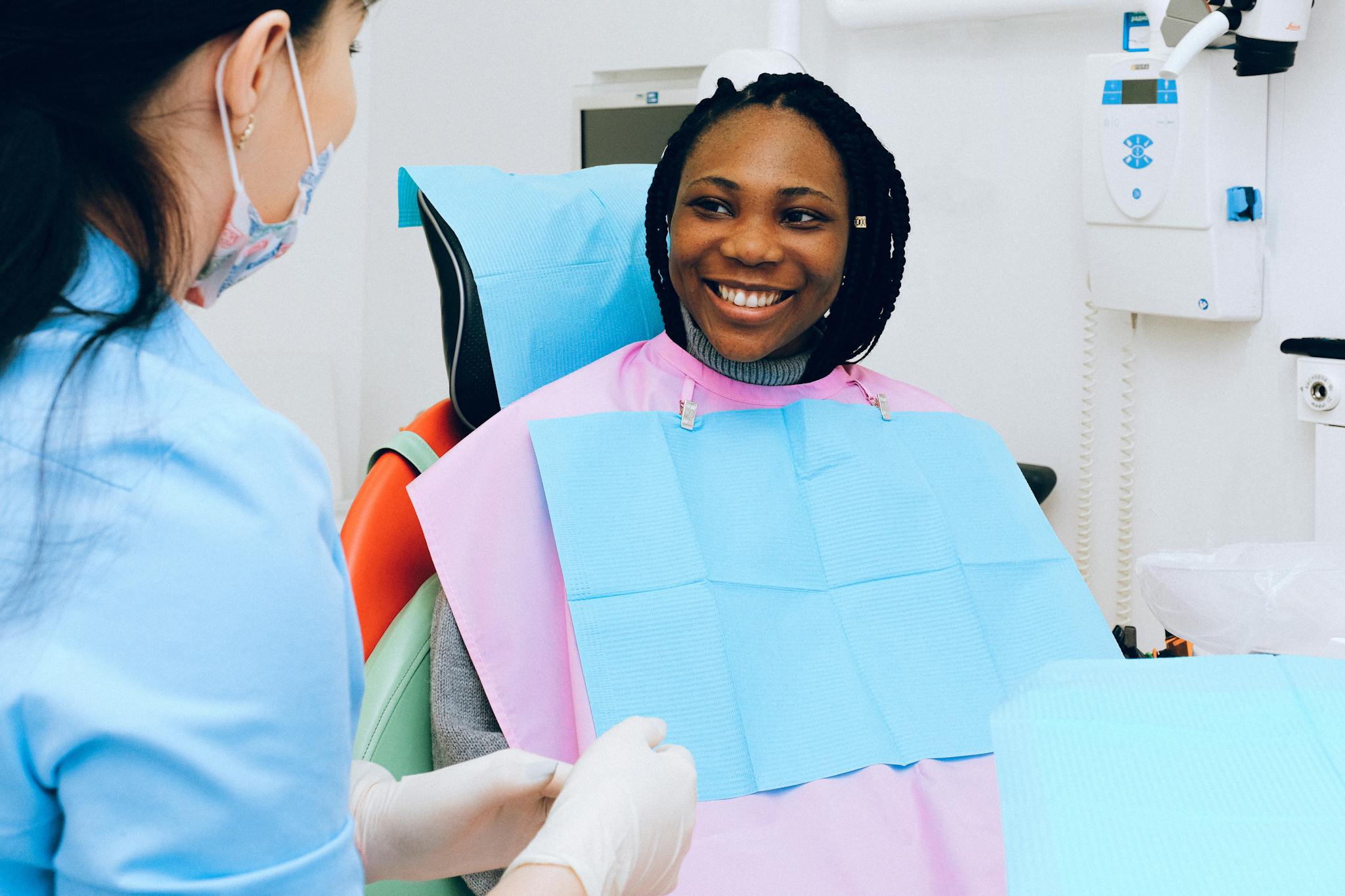Smiling woman receiving a dental check-up at The Village Dental, wearing a protective bib, with a dental professional in a blue uniform discussing oral health in a calm, professional setting.