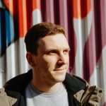 Man with a warm smile standing against a colorful background, representing patient satisfaction and the transformative cosmetic dental services at The Village Dental in Miami.